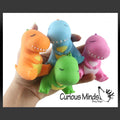 Mano sosteniendo cuatro juguetes squish de dinosaurios coloridos de PartyStore en naranja, azul, verde y rosa